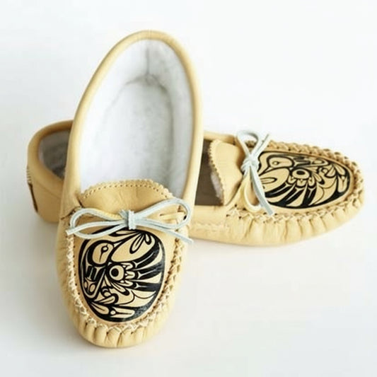 A pair of beige moccasins with black hummingbird artwork and light beige moosehide collar, featuring laces and set against a wooden floor background.