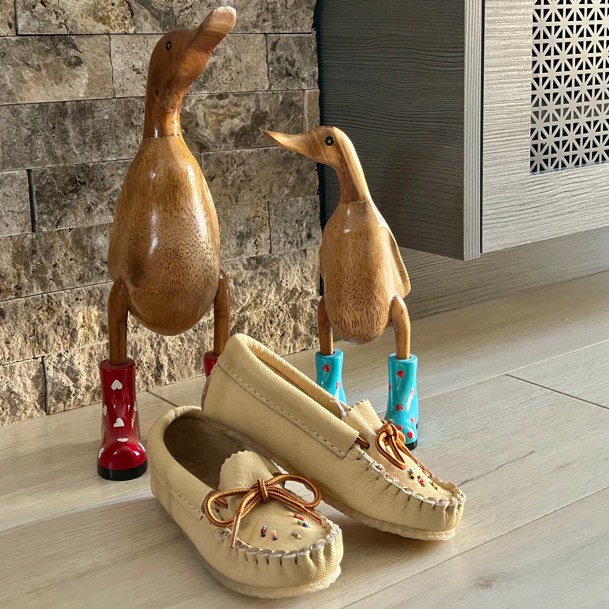 Beige moccasins with decorative elements on a wooden floor with two duck-shaped objects in the background.