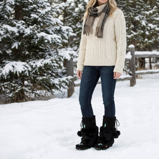 Person wearing a cream sweater, dark jeans, and black boots with pom-poms in a snowy landscape.