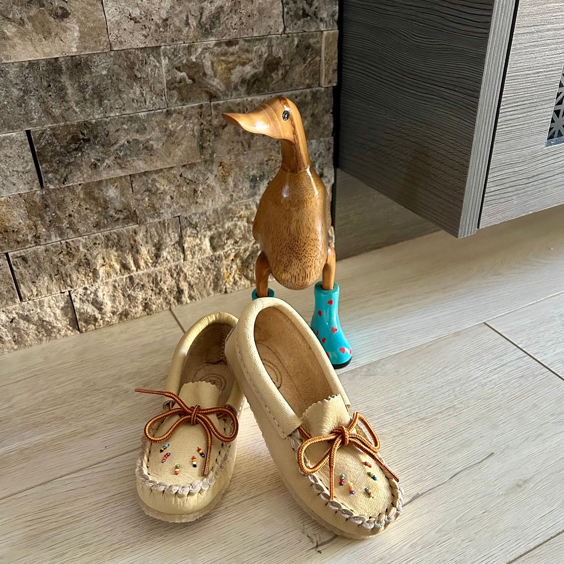 Pair of beige moccasins with decorative bows on a wooden floor, next to a decorative duck figure wearing blue boots.