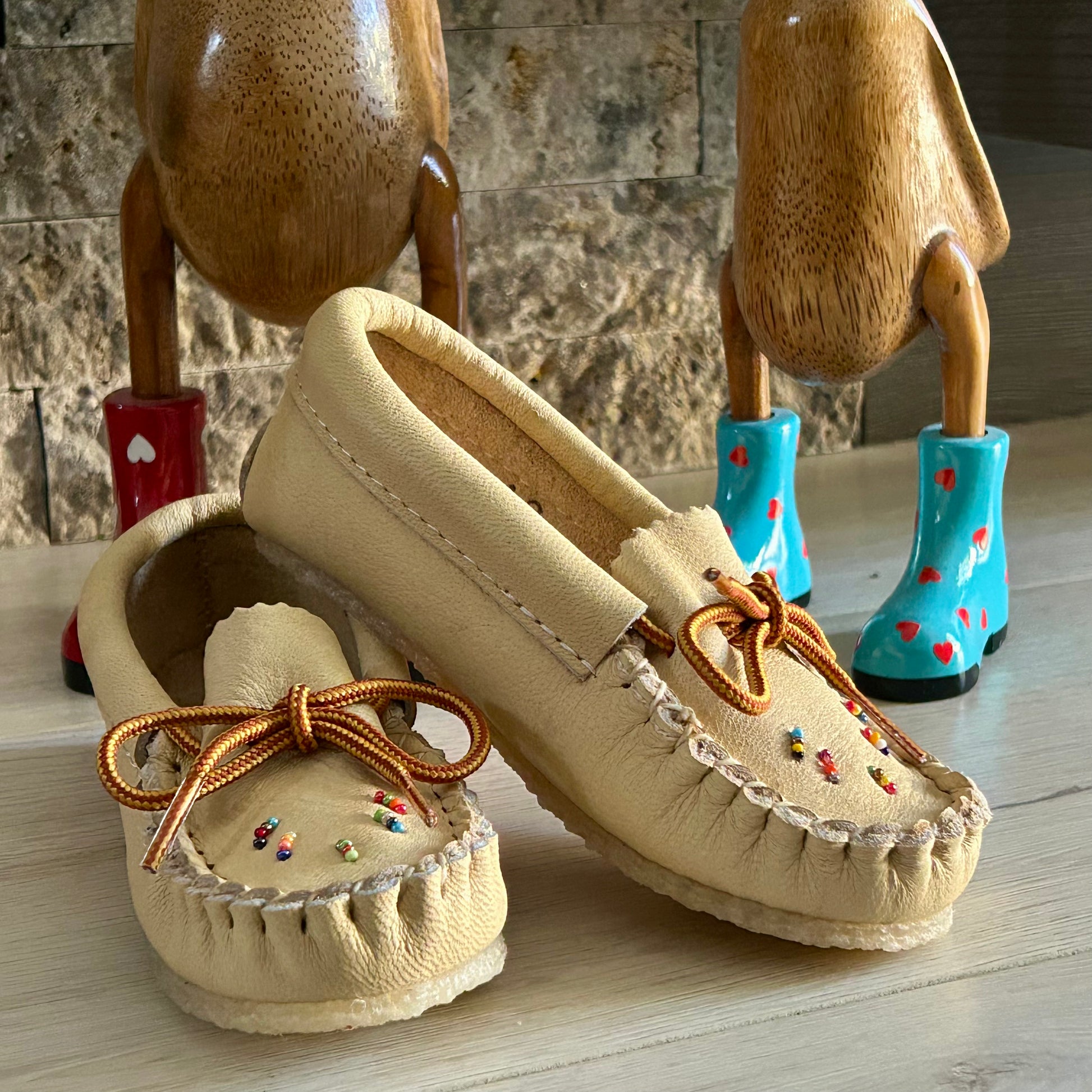 Pair of beige moccasins with brown laces on a wooden surface, with wooden figures and blue boots in the background.