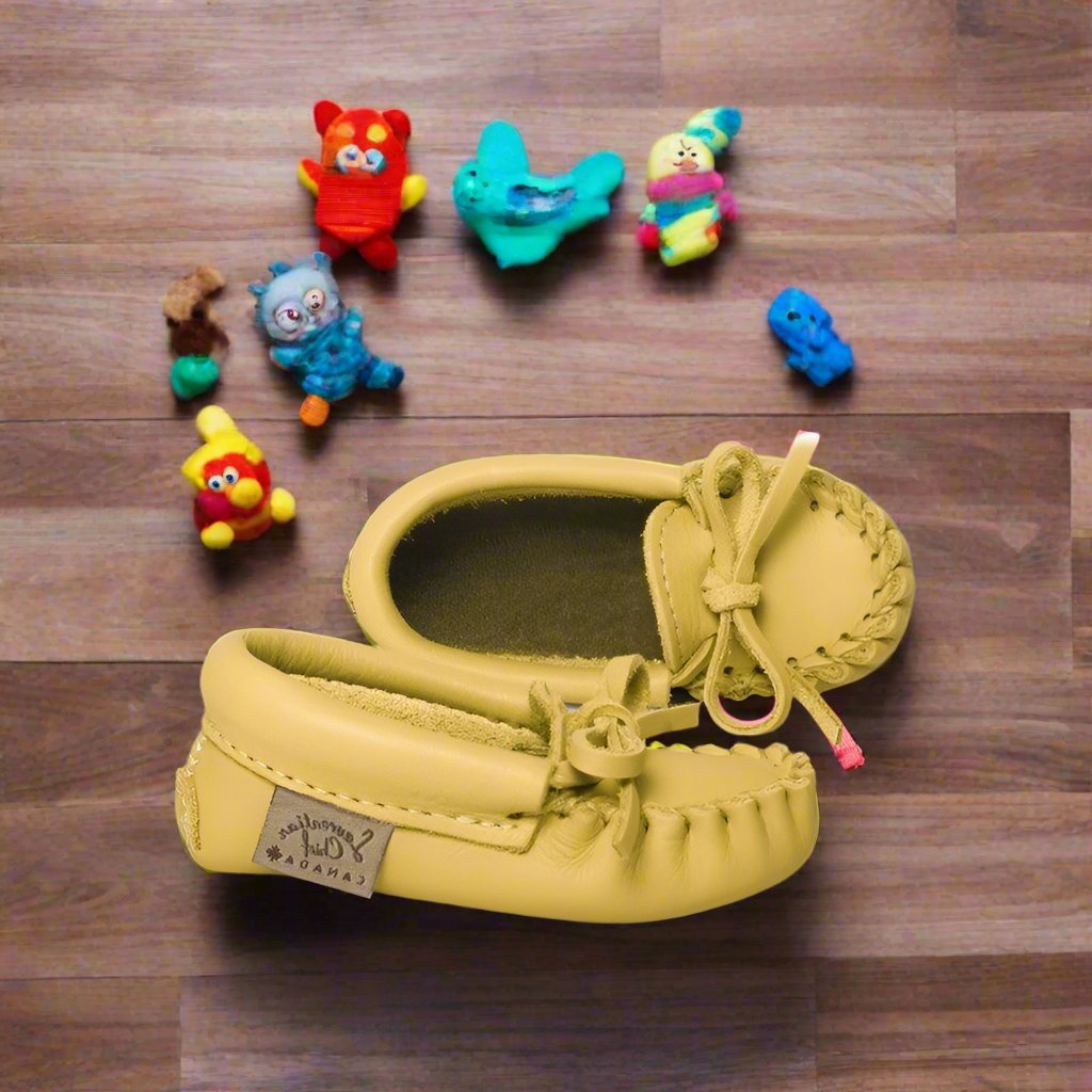 A pair of children's moosehide moccasins in yellow with laces and a brand label attached to the sole.
