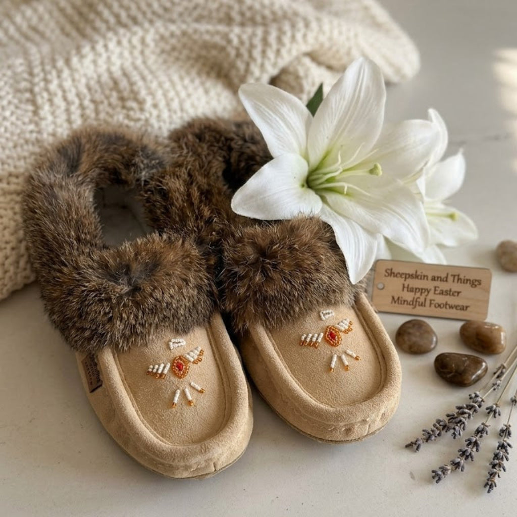 Pair of brown moccasin slippers with fur trim on a light surface with a white flower and stones.