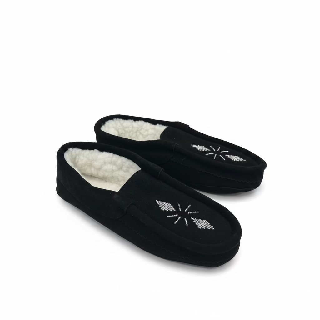 A pair of black suede moccasin-style slippers with white sherpa lining and silver snowflake embellishments, resting on a white tabletop.