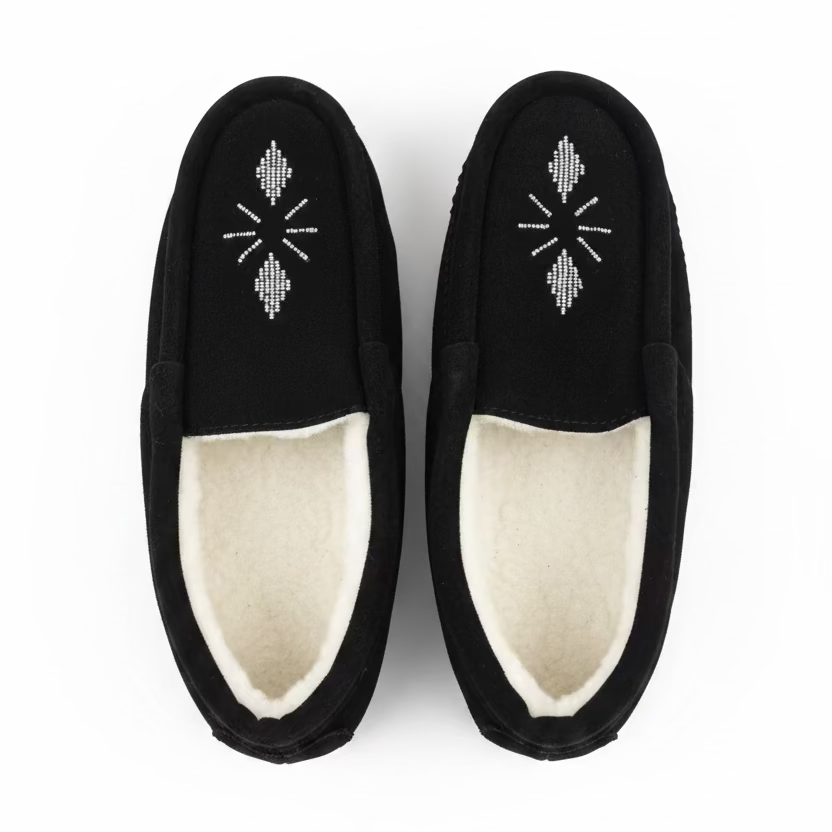 A pair of black slip-on slippers with white-sheaerling lining and a white geometric snowflake-like embroidery on the top, displayed on a light marble surface.