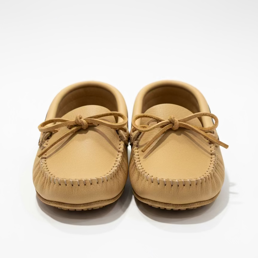 Small tan shoe with decorative band on a white surface, with a clock and plant in the background.